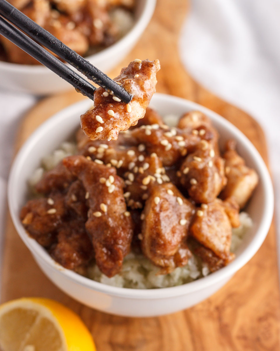 Chopsticks holding a bite od Chinese lemon chicken above a bowl.