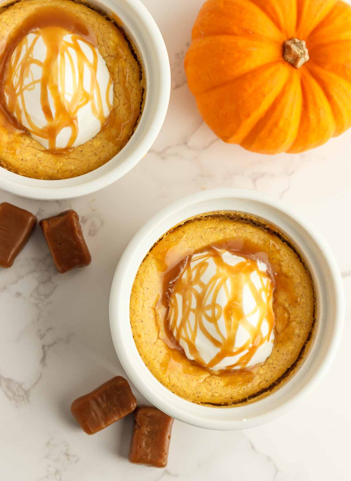 Two easy kettopumpkin cheesecakes in white ramekins with whipped cream and caramel syrup.