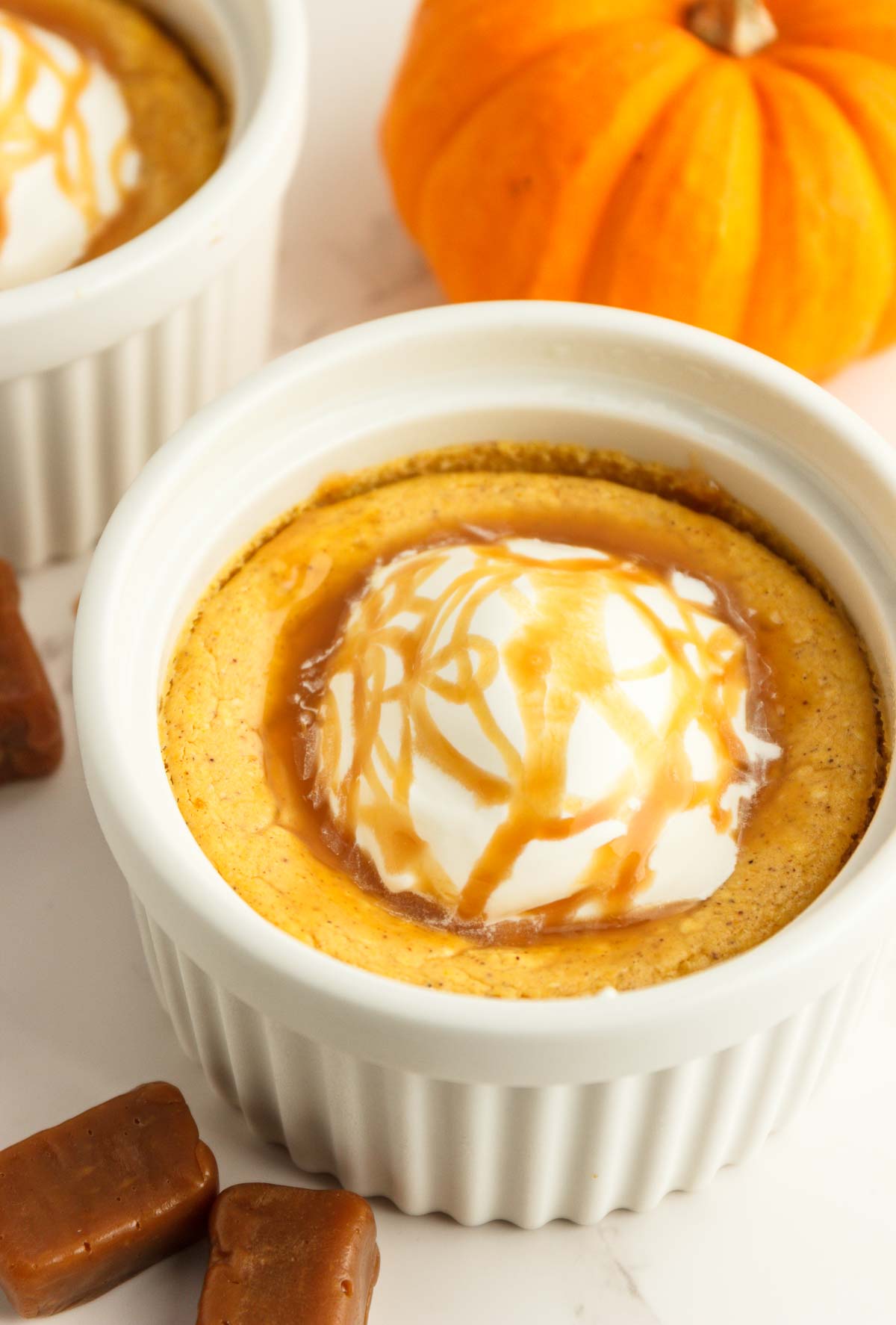 Two easy keto pumpkin cheesecakes in white ramekins with whipped cream and caramel syrup.